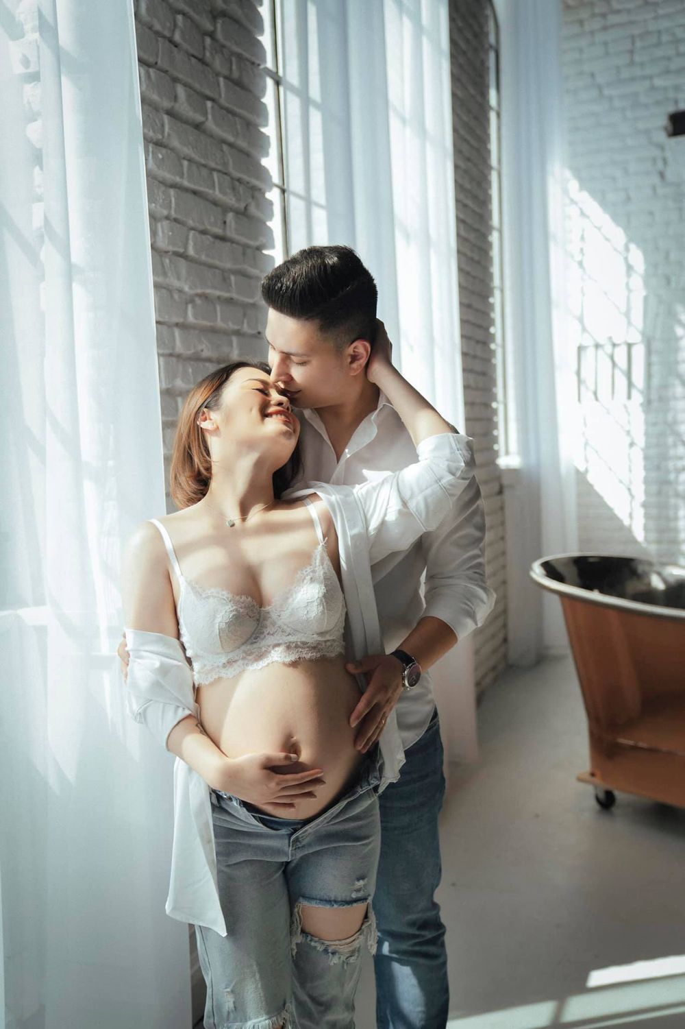 Intimate maternity photoshoot near a window with soft natural lighting.