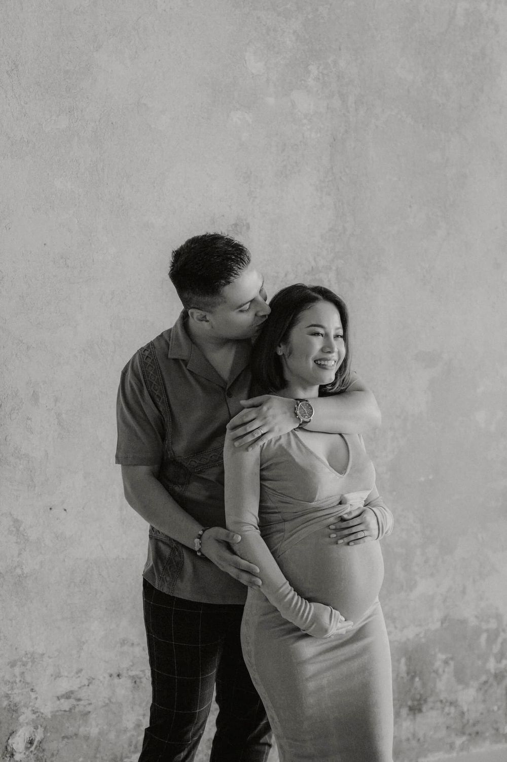 Black and white maternity photo showing a tender embrace against a textured wall.