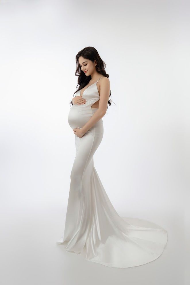 Expectant figure in white flowing gown poses against white backdrop in elegant maternity photo.