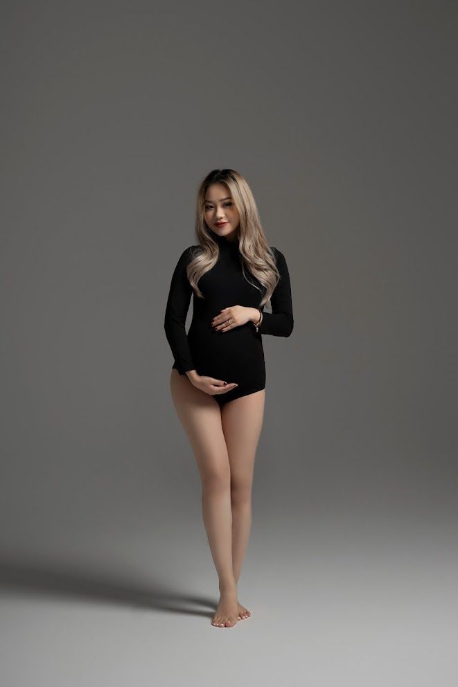 Elegant full-length maternity pose in black bodysuit against grey backdrop.