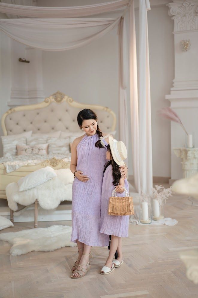 Two figures in matching purple dresses share a tender moment in a luxurious white bedroom setting.