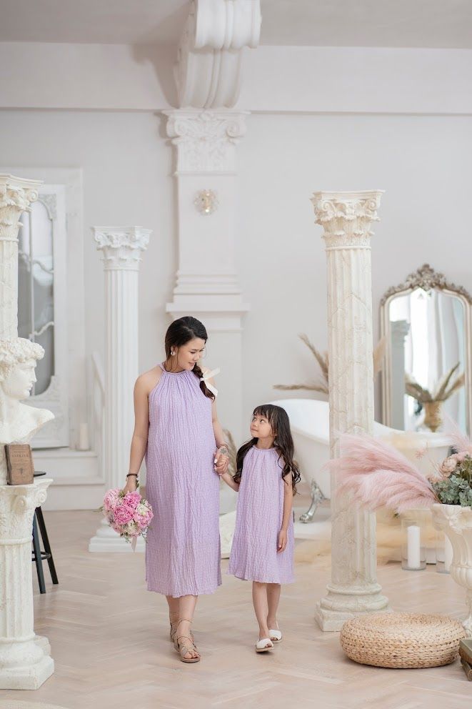 Two figures in matching lavender dresses pose together in an elegant white room with ornate columns and mirrors.