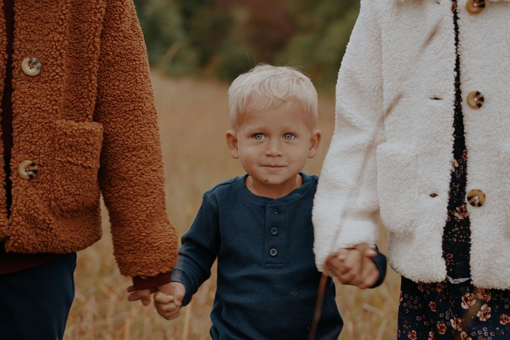 Young blonde toddler boy in navy shirt holding hands with two adults in an autumn field.