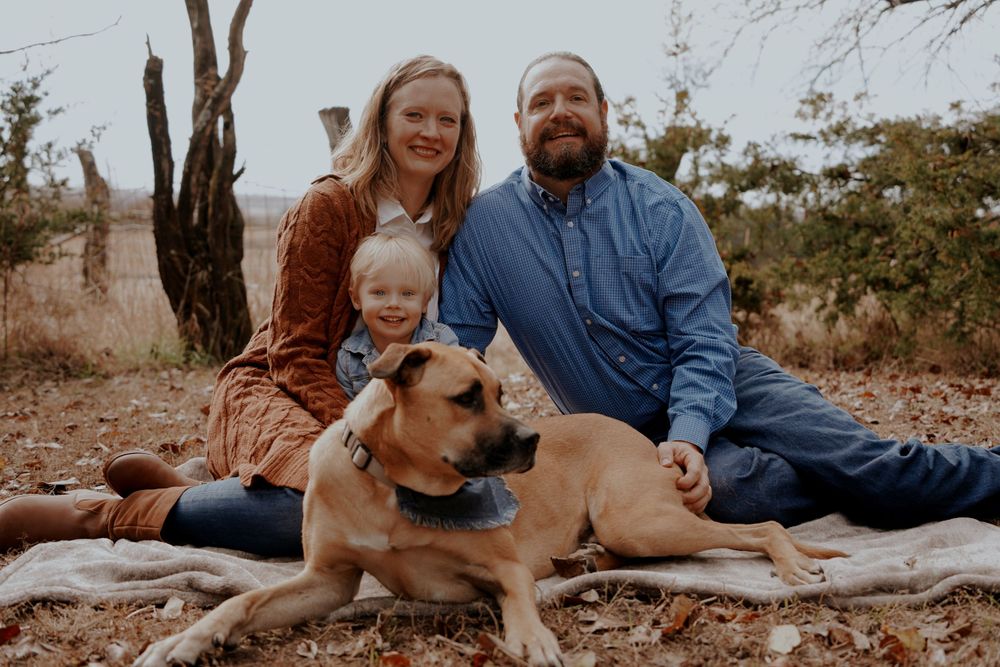 Family portrait with woman, man, and toddler sitting outdoors with a tan dog on a blanket among autumn trees.