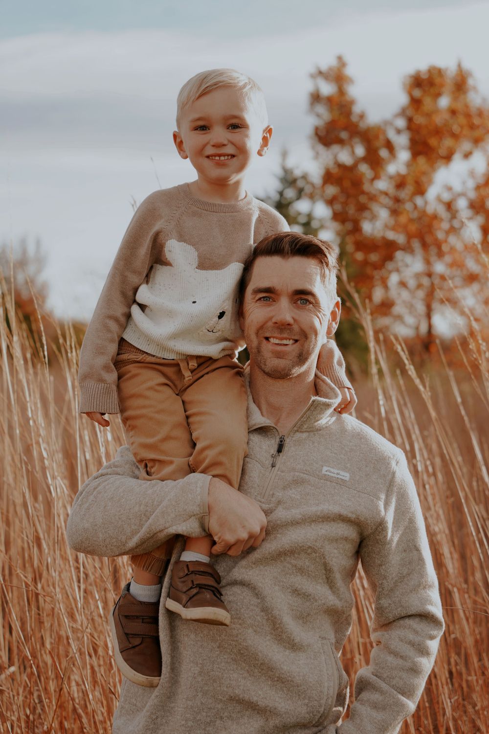 Smiling father carrying young son on his shoulders in a tall golden autumn grass field.