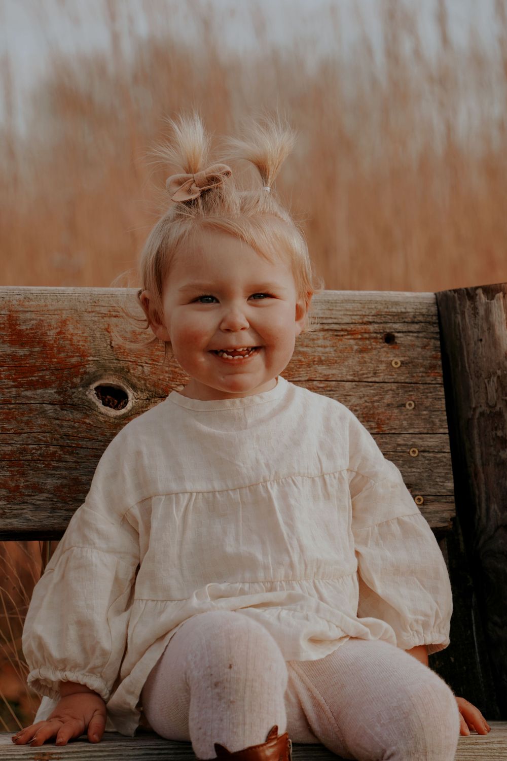 Smiling toddler girl with pigtails wearing a white dress sitting on a wooden bench outdoors.