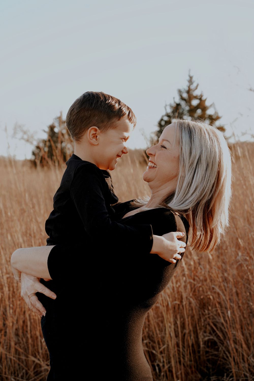 Mother in black outfit holding her young son in a golden field, sharing a joyful moment.