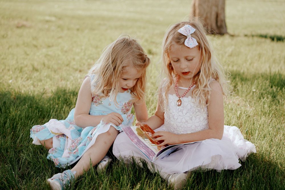 Two young blonde girls in dresses sitting on green grass looking at a colorful book together.
