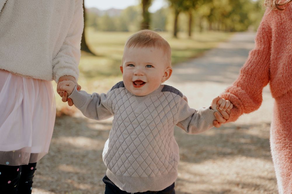 Happy toddler boy in a grey sweater laughing while holding hands with adults on a sunny path.