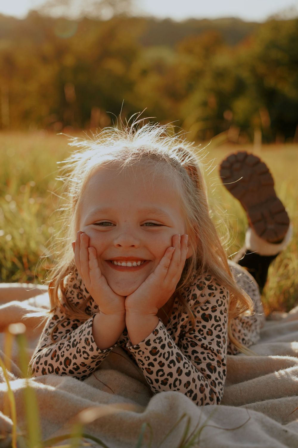 Young blonde girl lying on a blanket in a sunny field, smiling with hands on her cheeks.