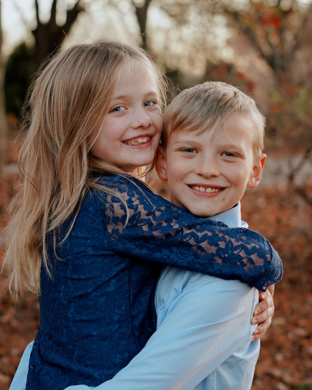Smiling girl in blue lace dress hugging a boy in a light blue shirt among autumn leaves.