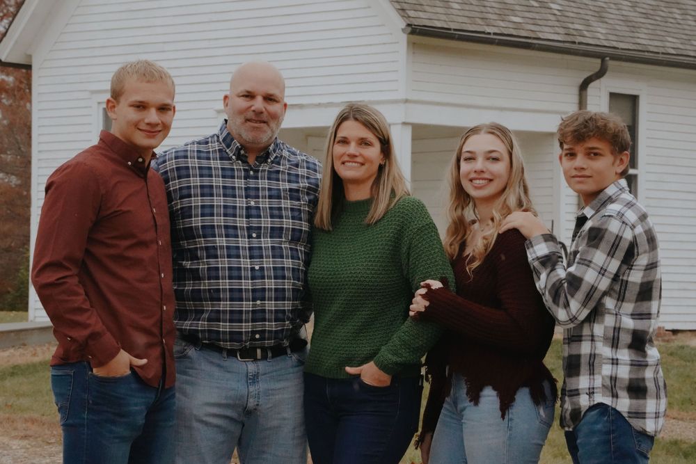 Family of five smiling together outdoors near a white building in autumn casual attire.