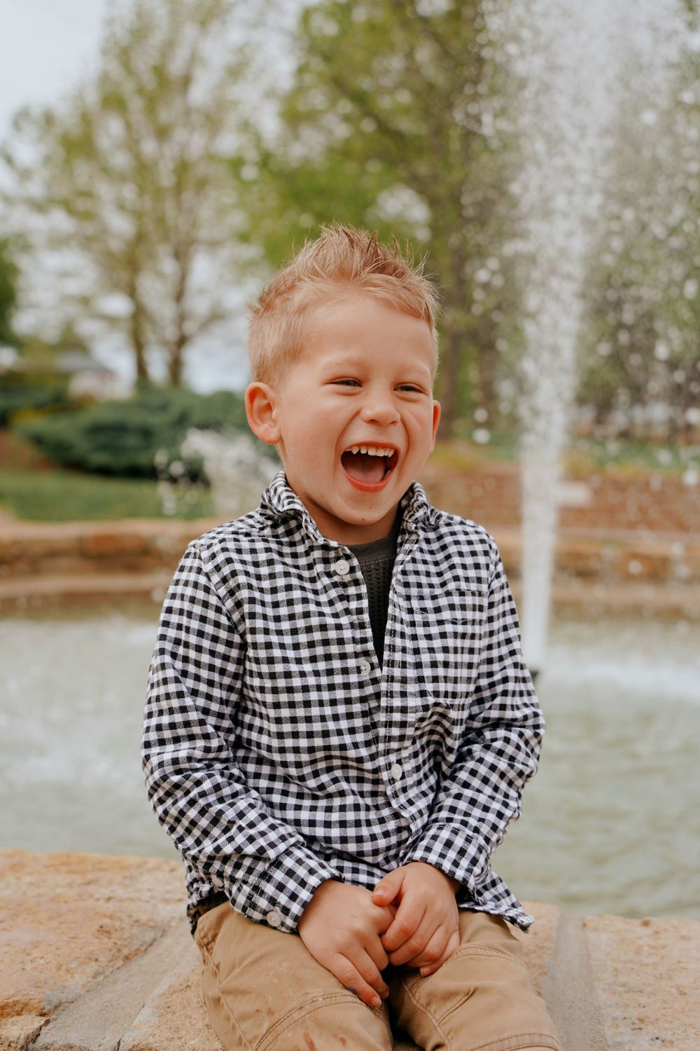 Laughing young boy in a plaid shirt sitting near a fountain in a park setting.