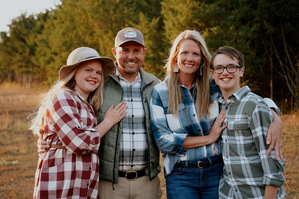 A happy family of four poses together in a sunlit field wearing coordinating flannel shirts for fall portraits.
