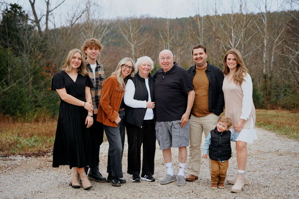 Multi-generational family of seven smiling together on a gravel path surrounded by fall foliage.