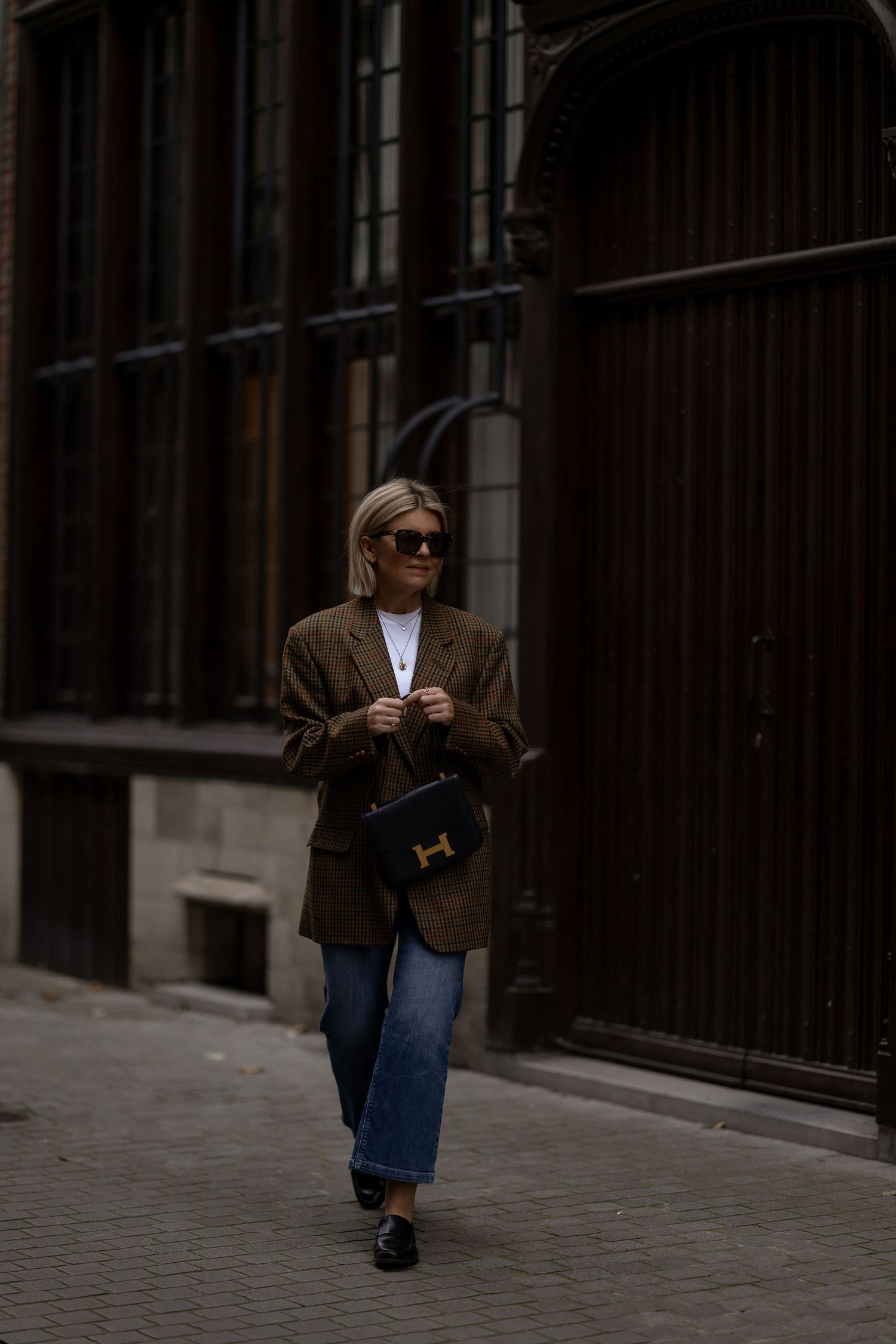 Stylish street fashion look featuring brown blazer, blue jeans and black Hermes bag on city sidewalk.