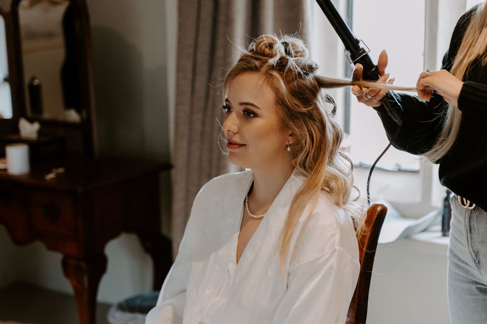 Bride getting her hair done on her wedding morning. she is wearing a white satin dressing gown.