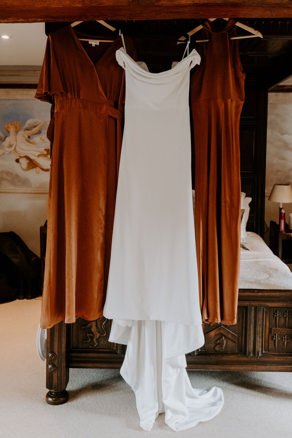 an ivory wedding gown and 2 burnt orange bridesmaid dresses hanging from a four poster bed.