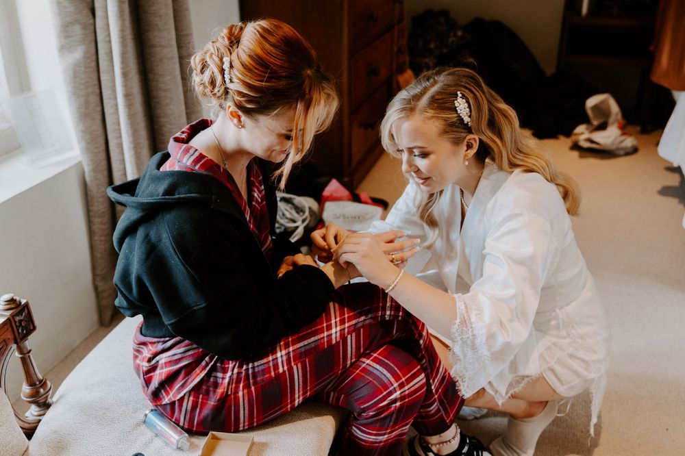 a bride doing up a bracelet on her bridesmaid. the bridesmaid is sat on a chaise lounge and the bride is kneeling beside her.