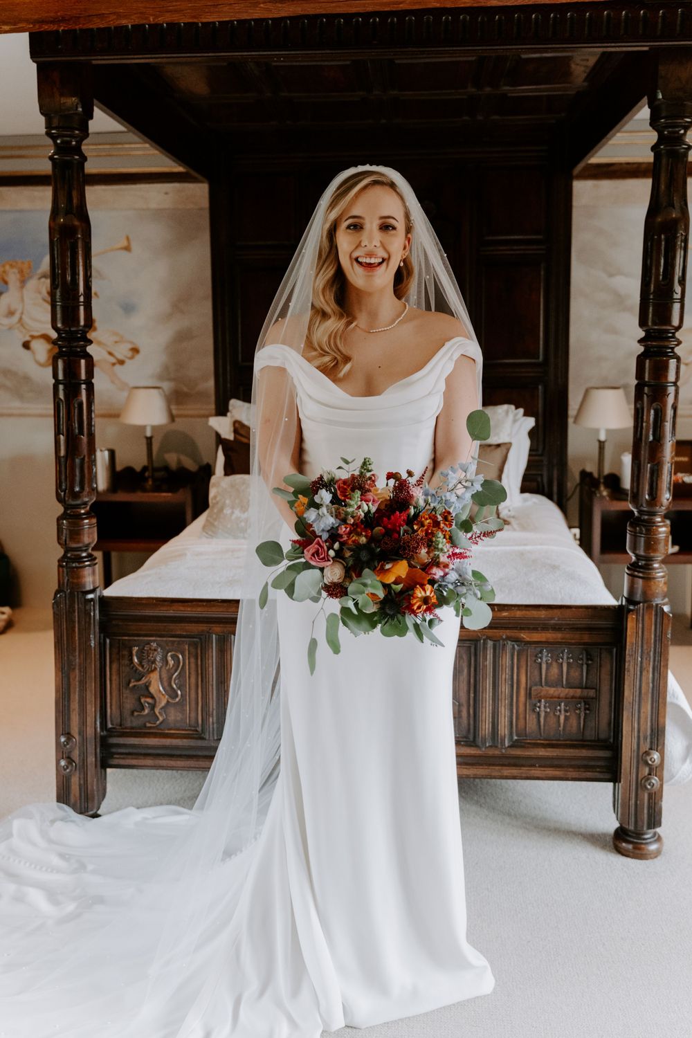 a bride in an ivory, figure hugging gown and a long veil. she is holding a autumnal, bright bouquet and is laughing towards the camera.