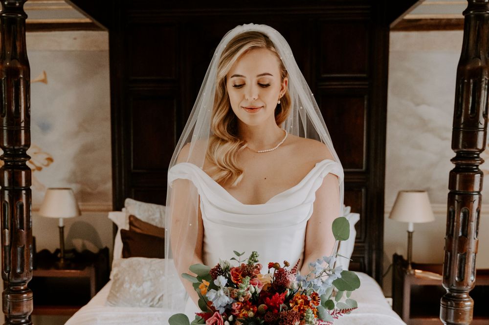 a bride in an ivory, figure hugging gown and a long veil. she is holding a autumnal, bright bouquet. bride is looking down at her flowers