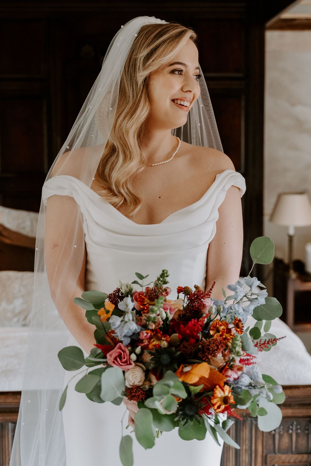 a bride in an ivory, figure hugging gown and a veil. she is holding a autumnal, bright bouquet. Bride is looking to her left and smiling.