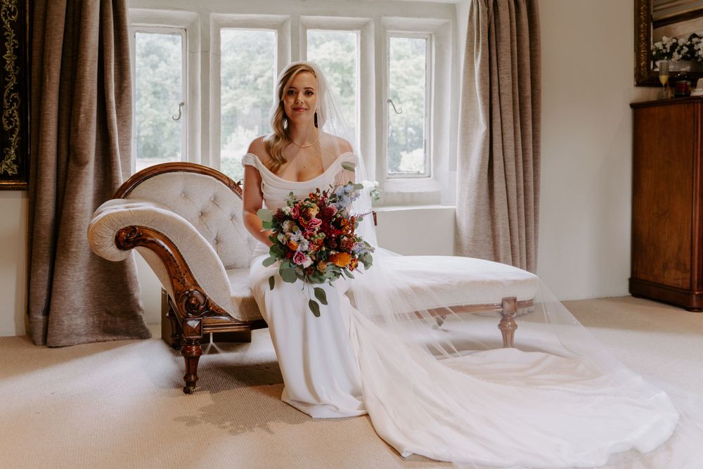 a bride is sat on a chaise lounge in an ivory, figure hugging gown and a long veil. she is holding a autumnal, bright bouquet.