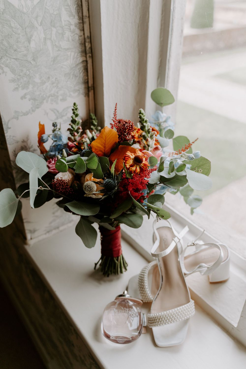 a bright colourful wedding bouquet sat on a window sill. Bedside them is a pair of ivory scrappy wedding shoes.