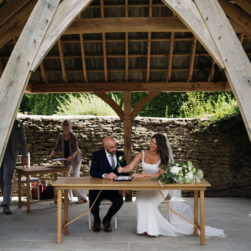 Newlyweds sit at a wooden table under a rustic stone arch during their wedding ceremony.