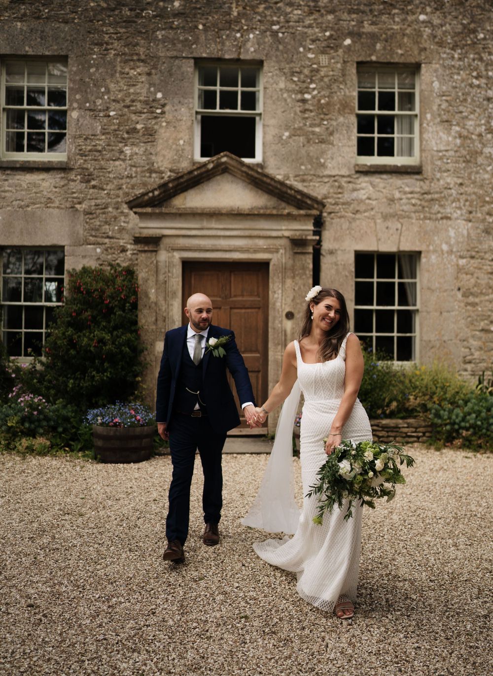 A couple poses on the gravel driveway in front of a historic stone manor house.