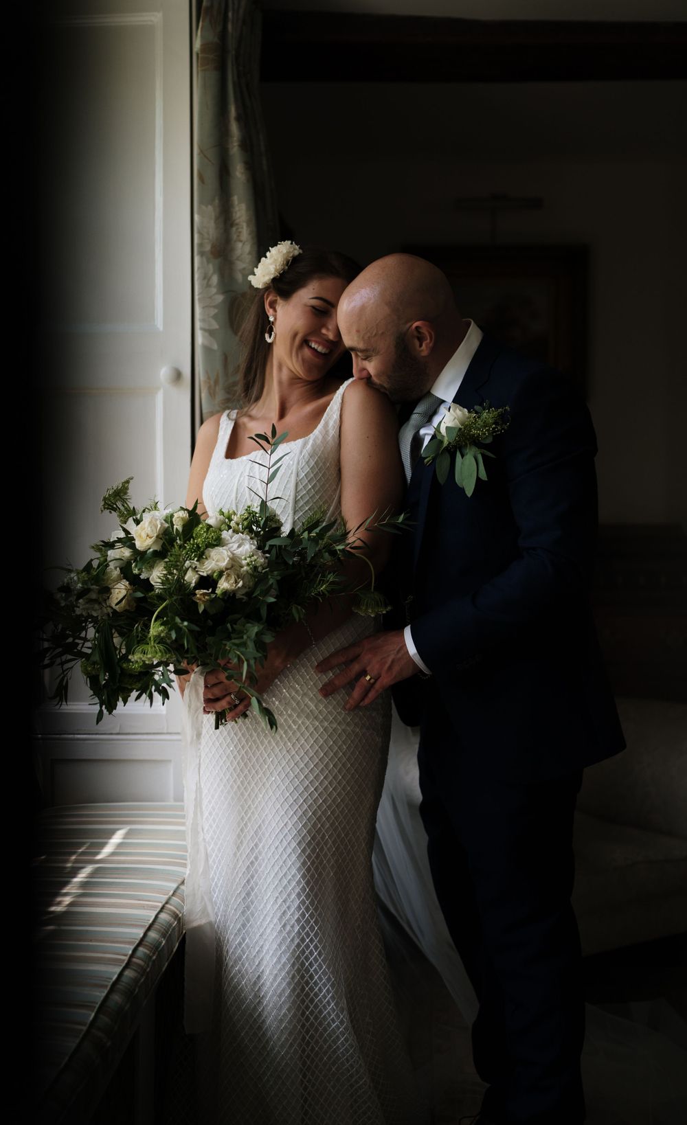 Intimate moment captured near a window with elegant white floral bouquet.