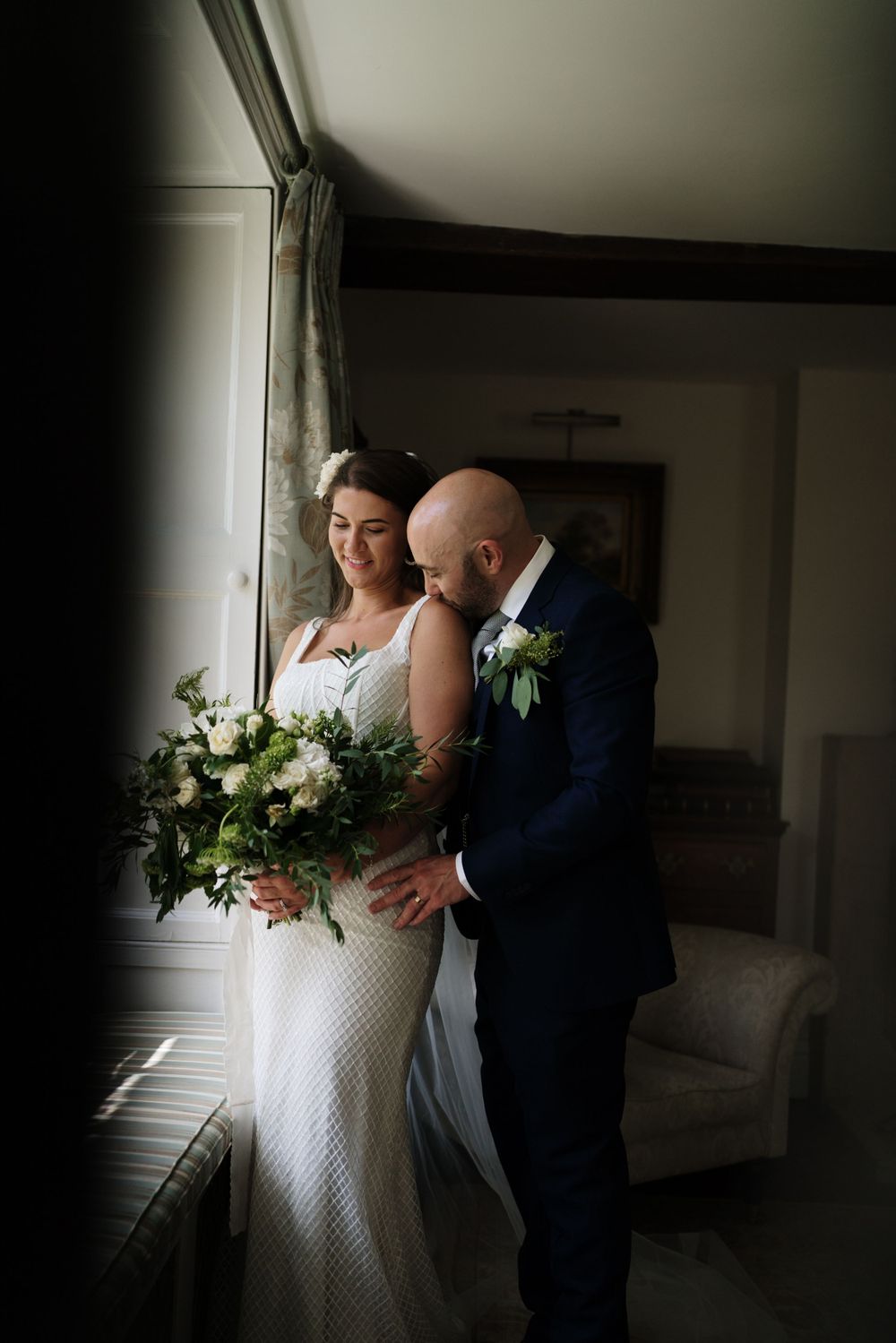 Moody window light illuminates a tender moment with greenery bouquet.
