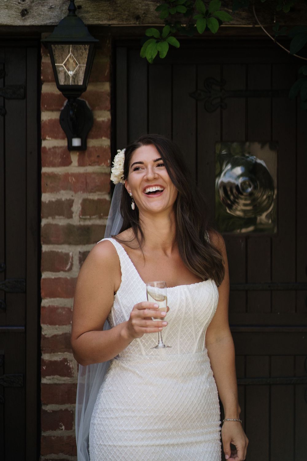 A joyful bride in a fitted lace gown holds a champagne glass while laughing.