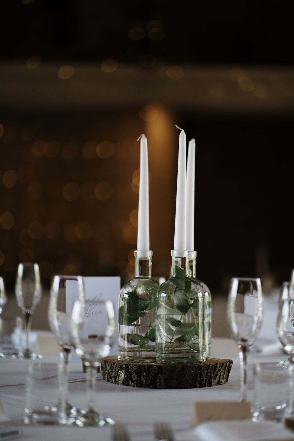 Elegant table setting with white candles in glass bottles on wooden slice centerpiece.