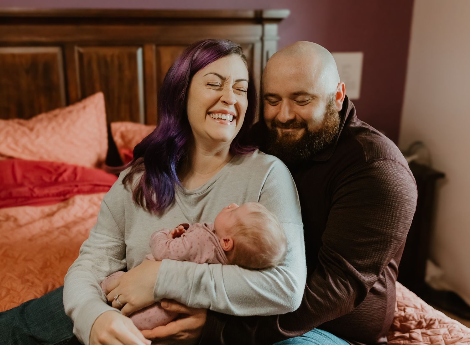 Happy parents share a joyful moment with their newborn on a bed with pink pillows.Colorado Springs Photographer