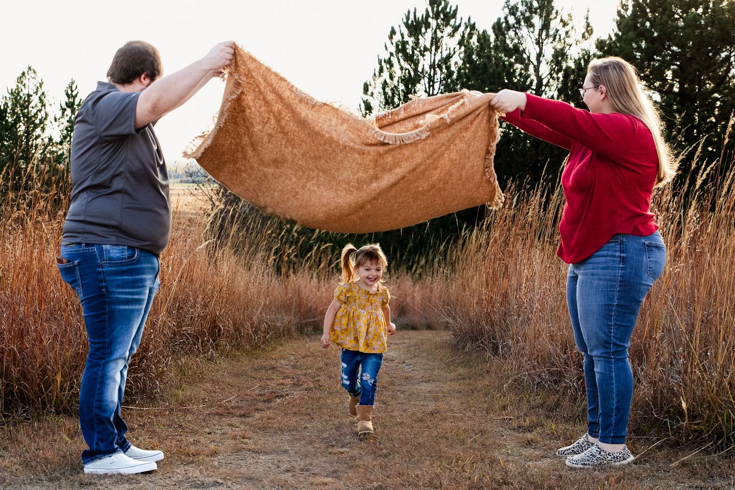 Two people holding a blanket up while a small child runs underneath on a nature path.