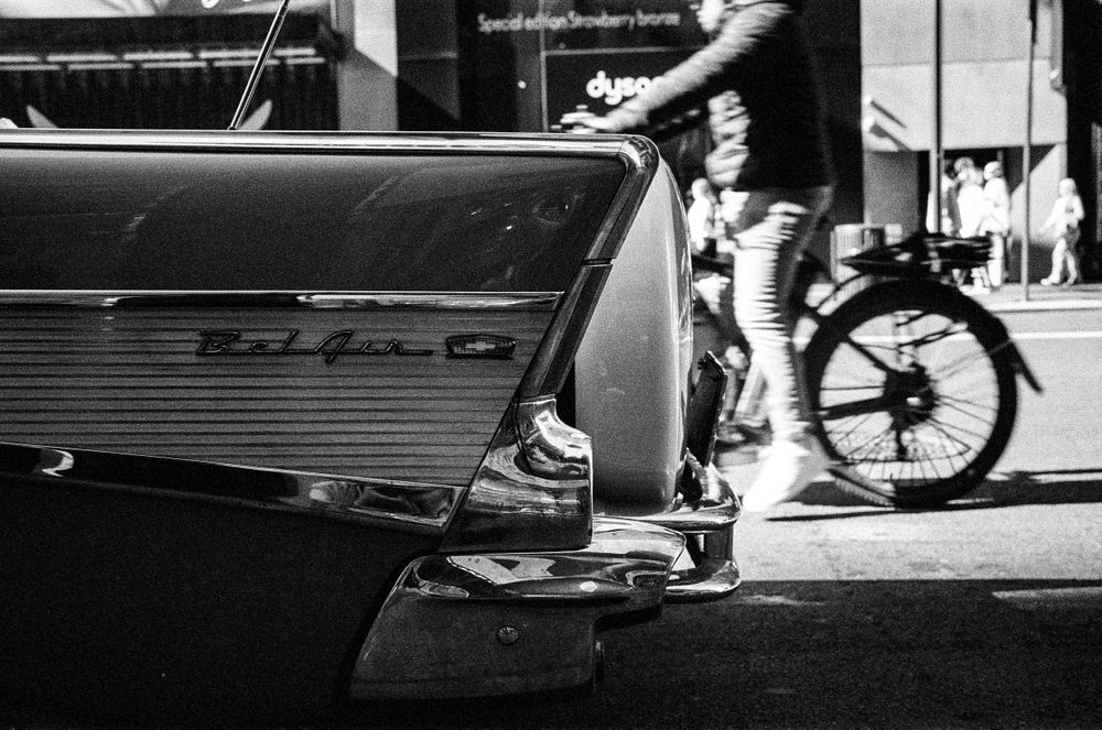Close-up of a vintage car's chrome trim with a cyclist passing in the background.