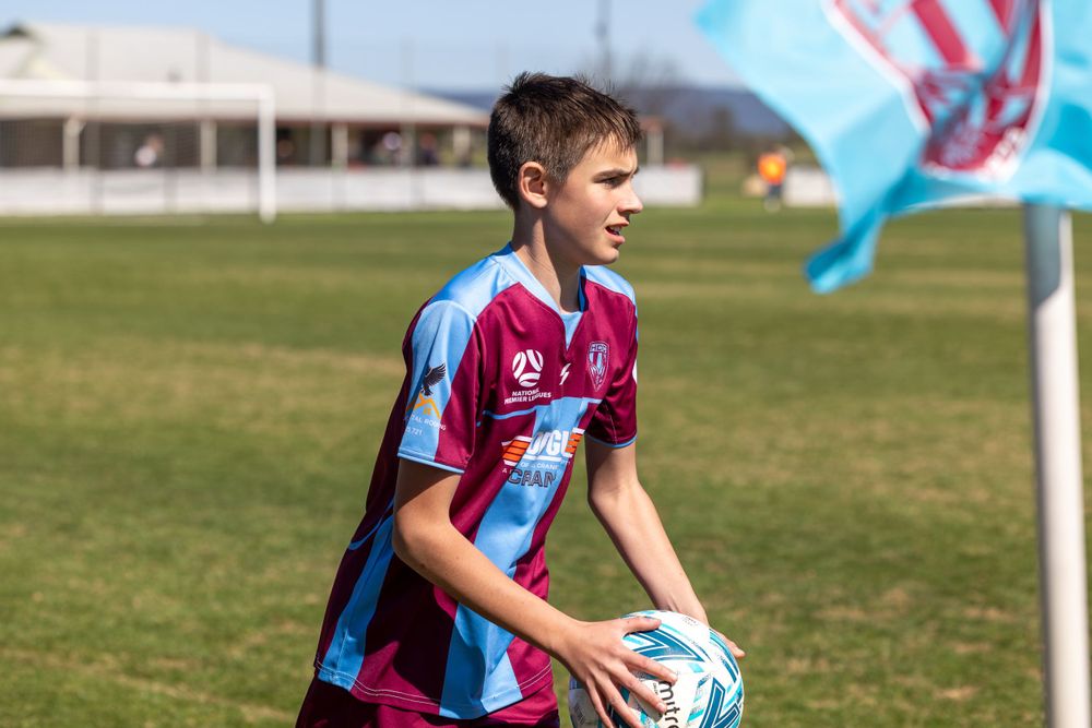 Young player in claret and blue striped jersey holds a soccer ball on sunny field.