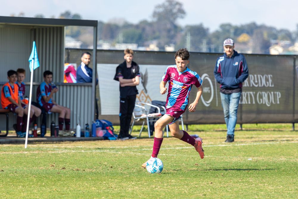 Soccer player in maroon and blue stripes kicks ball during match on grass field.