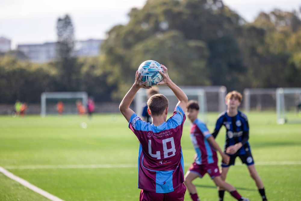 Soccer player in maroon and blue uniform throwing ball in during a match on a grass field.