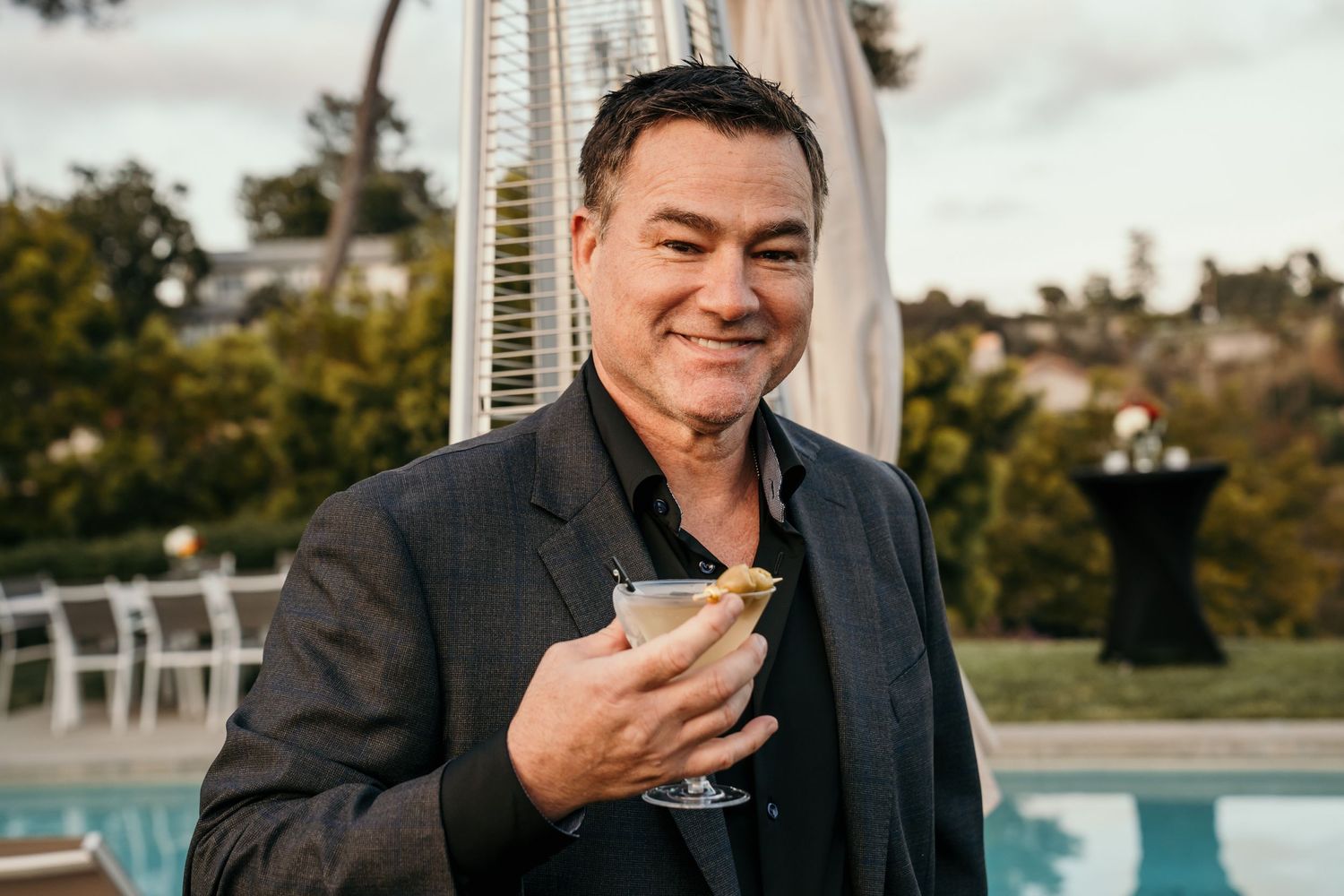 Man in dark blazer enjoying cocktail at outdoor poolside venue.