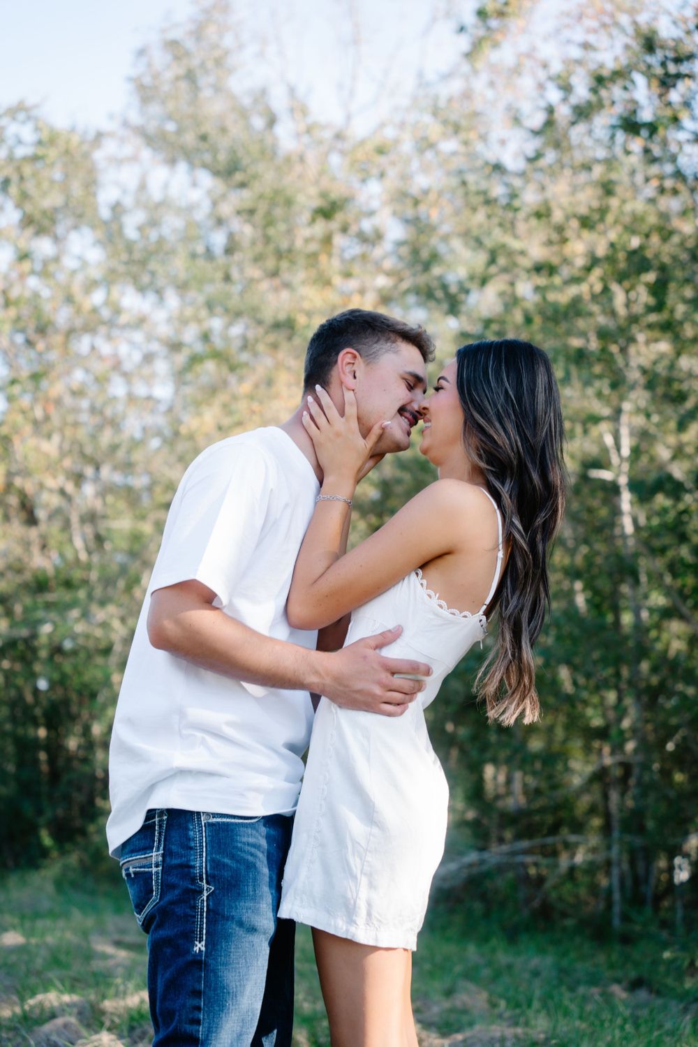 Romantic couple in white outfits embrace in wooded setting during golden hour.