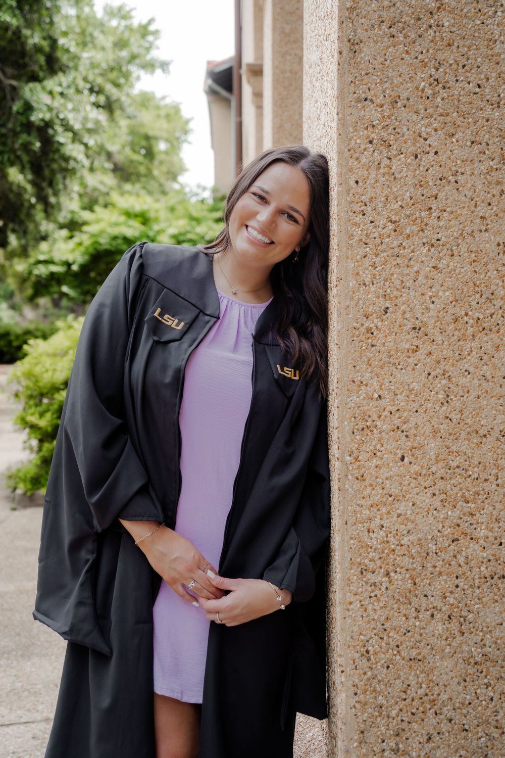 Graduate in black gown and purple dress poses against textured wall with natural lighting.