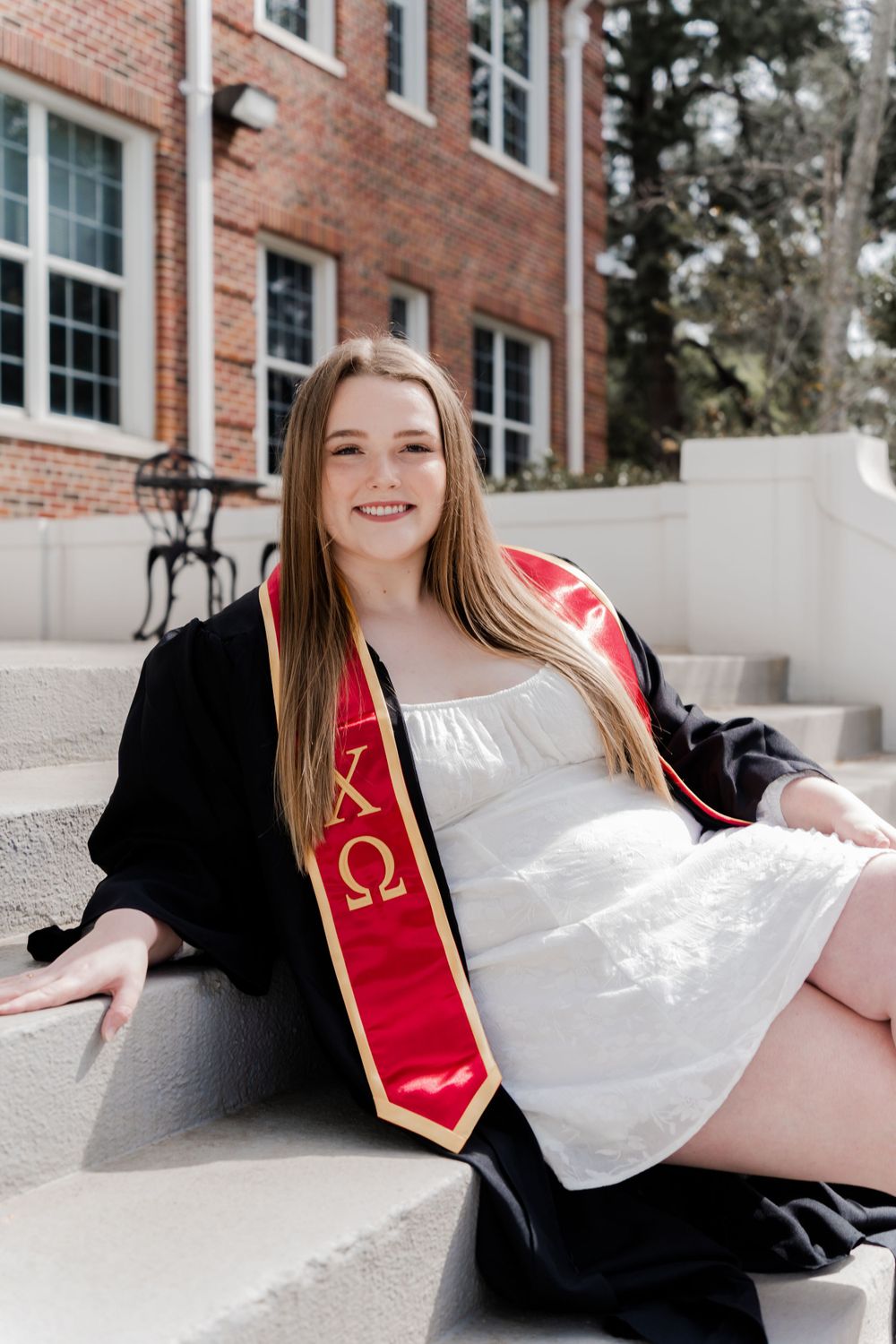 Graduate in white dress wearing Chi Omega stole sits on campus steps during graduation photoshoot.