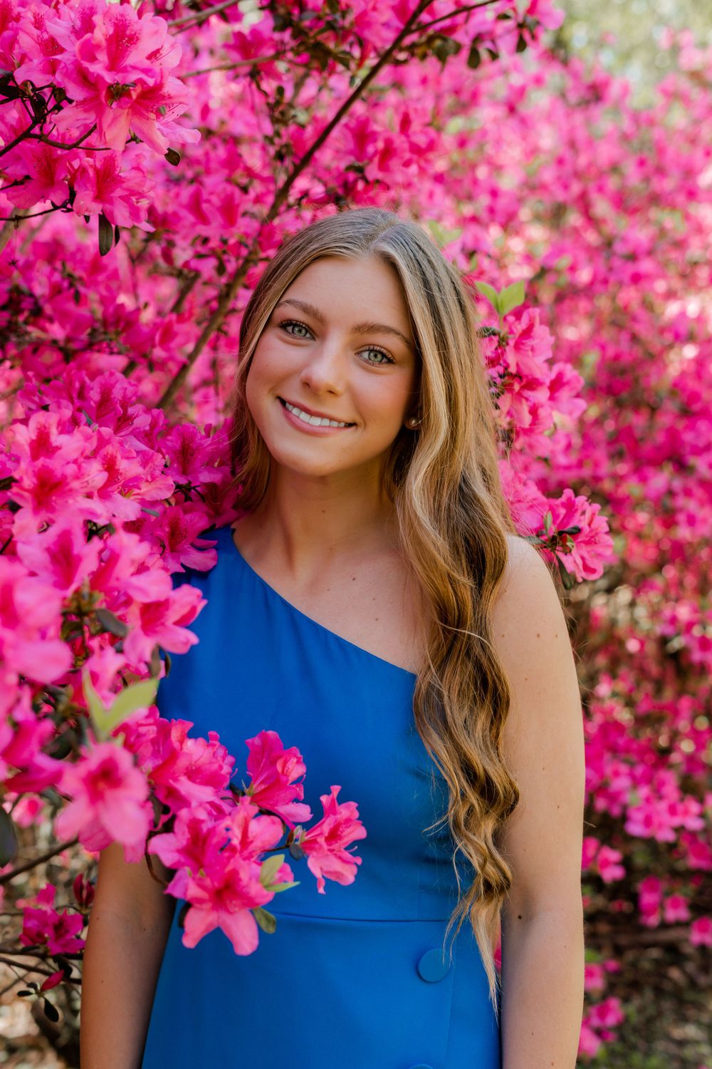 A person in a one-shoulder blue dress poses in front of vibrant pink azalea bushes in bloom.