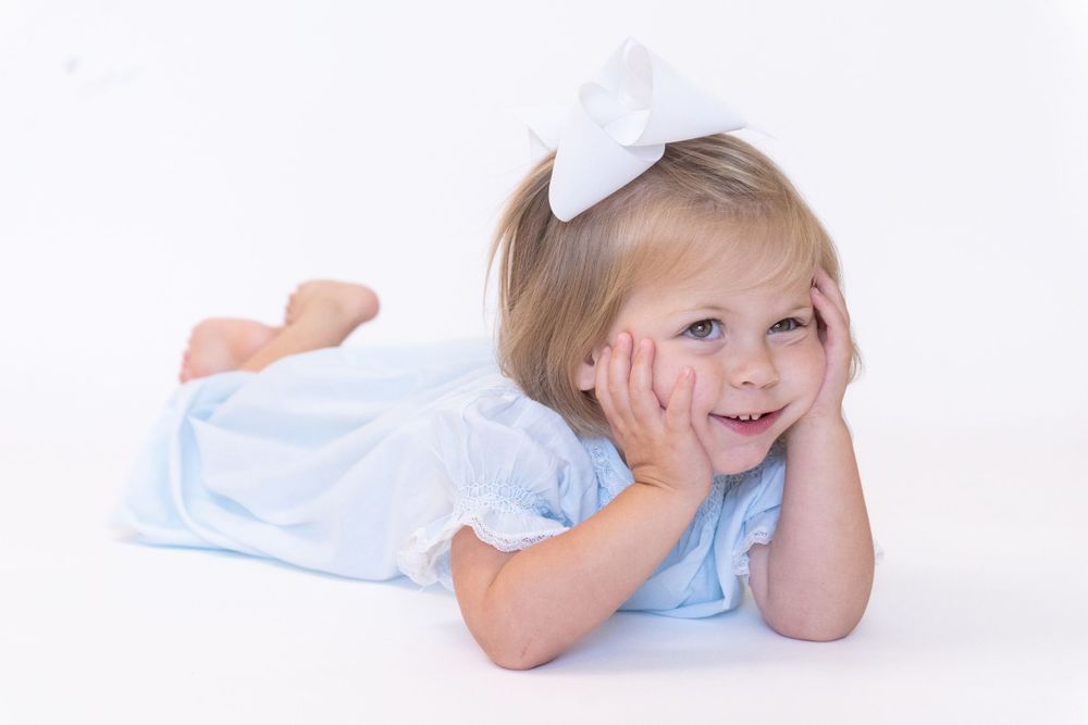 Child in light blue dress and white bow lying on white backdrop with cheerful smile.
