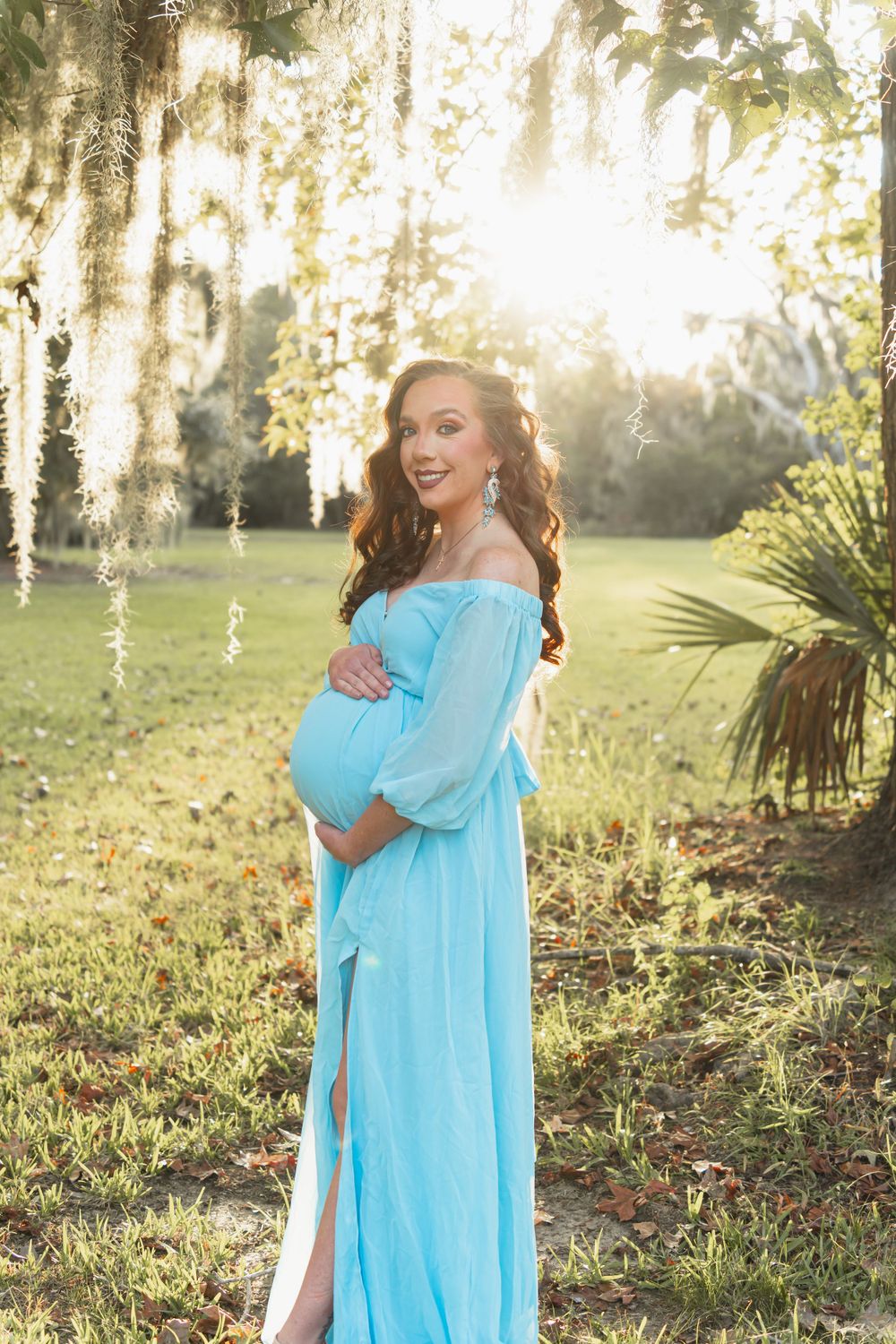 Maternity portrait in flowing blue dress against sunlit garden with Spanish moss.