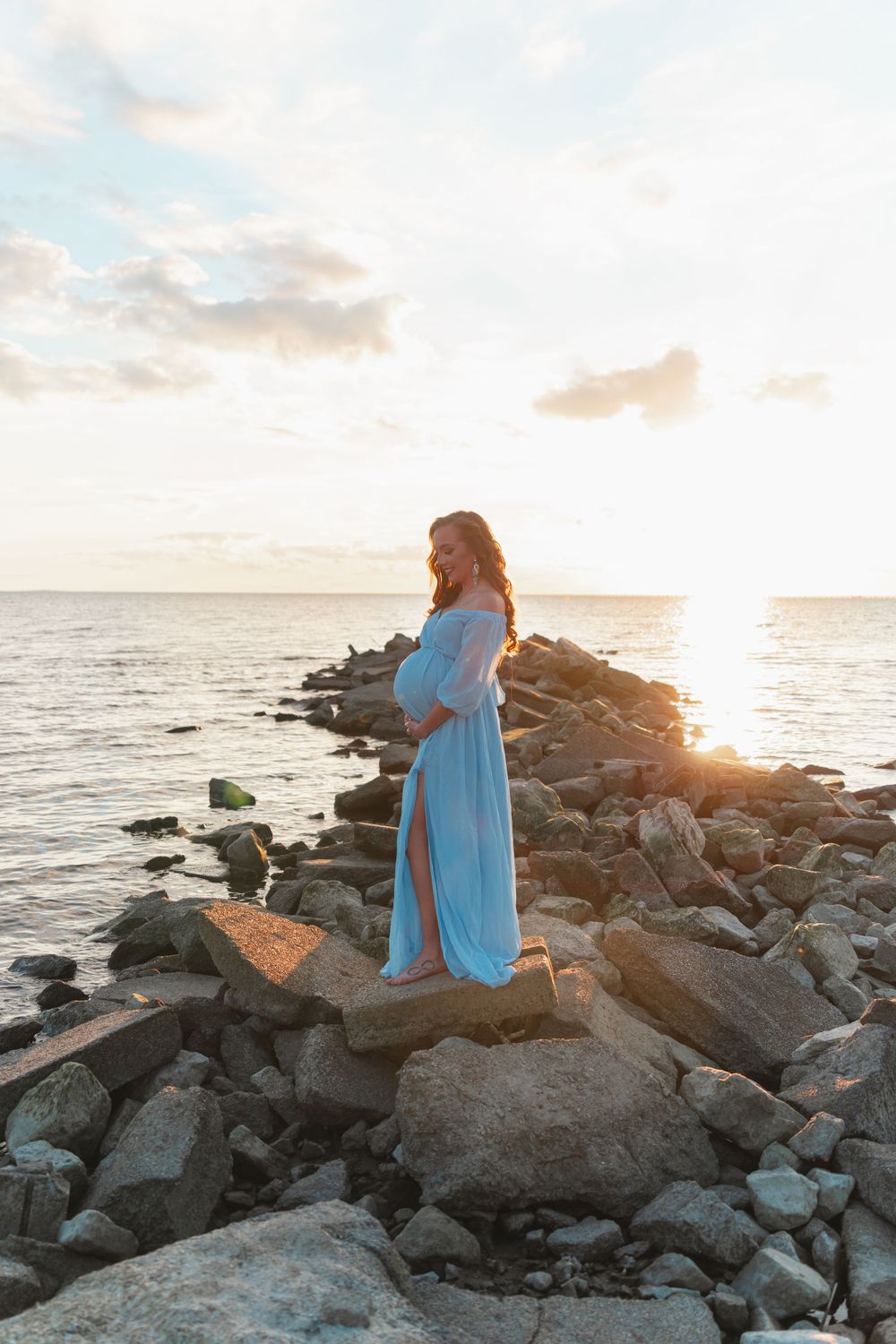Woman in flowing blue dress stands on rocky coastal outcrop at sunset by the ocean.