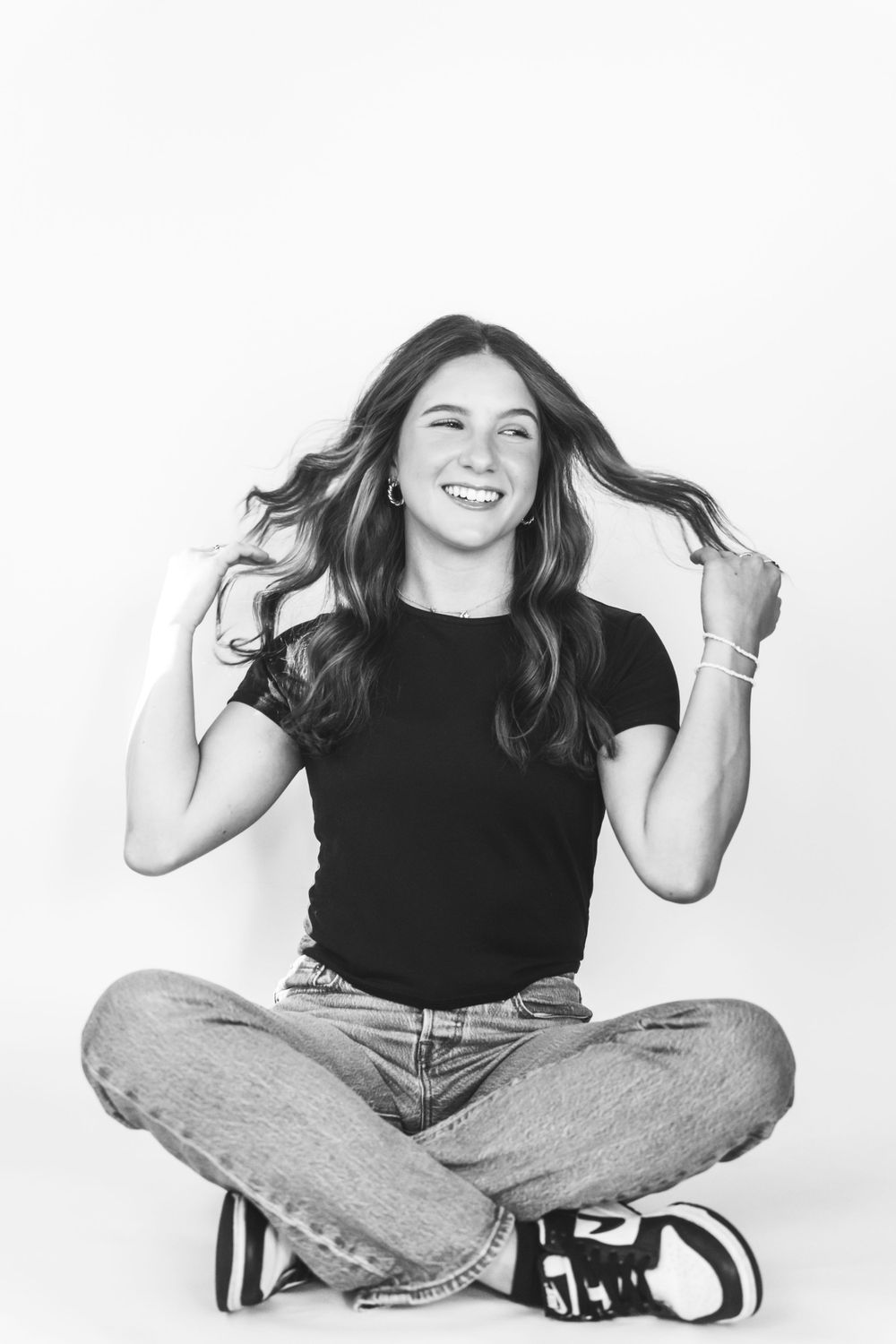 A joyful person in casual attire sits cross-legged on a white background while playing with their hair.