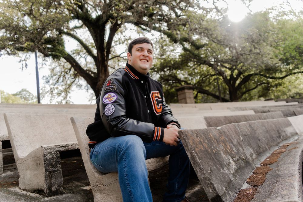 A person in a varsity jacket sits outdoors on stone steps surrounded by trees.